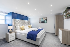 A modern bedroom with a tufted headboard, a white bed with blue bedding, a nightstand with drawers, a wooden dresser, and a wall-mounted shelf with plants.