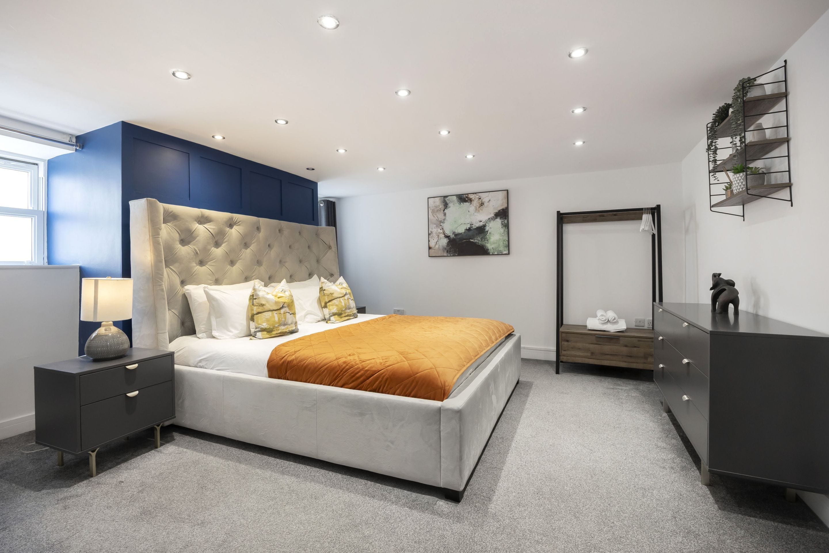 Un dormitorio moderno con una cabecera capitonada, una cama grande con colcha color mostaza, una mesita de noche con lámpara y un armario.