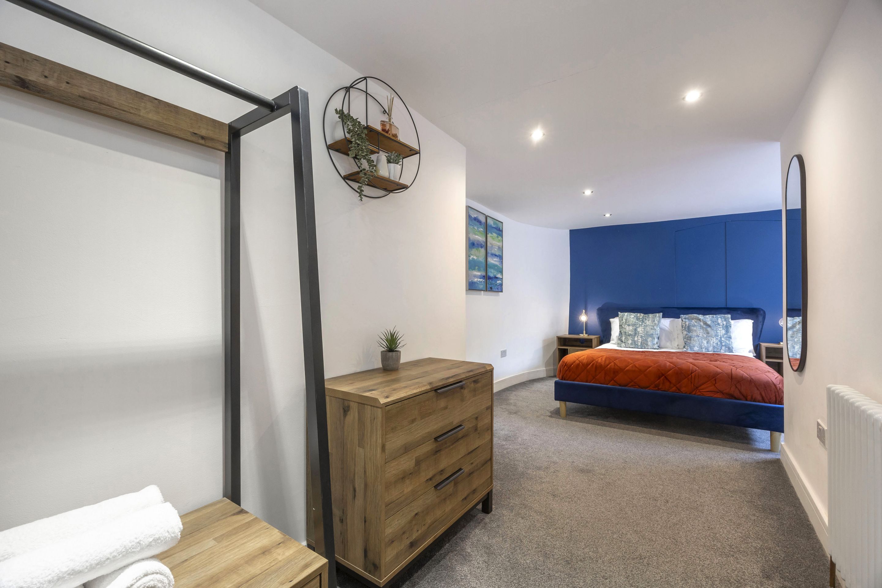 A bedroom with a bed, a wooden dresser, a mirror, and a wall-mounted shelf with plants.