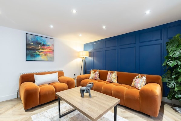 A modern living room with two orange sofas, a wooden coffee table, a colorful abstract painting, and a large green plant.