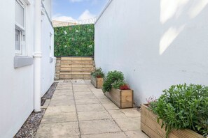 A narrow courtyard with a stone pathway, white walls, and wooden planters with greenery.