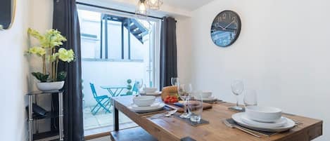 A dining area with a wooden table set for four, a black wall clock, and a potted plant.
