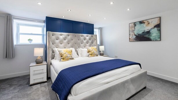 A modern bedroom with a tufted headboard, a large bed with a blue bedspread, two bedside lamps, and a wall-mounted abstract painting. - Large Period 2-Bed Apartment Close to Town (Gloucestershire)