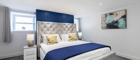 A modern bedroom with a tufted headboard, a large bed with a blue bedspread, two bedside lamps, and a wall-mounted abstract painting.