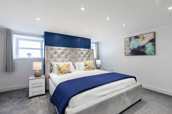 Un dormitorio moderno con una cabecera capitonada, una cama grande con colcha azul, dos lámparas de noche y un cuadro abstracto colgado en la pared.