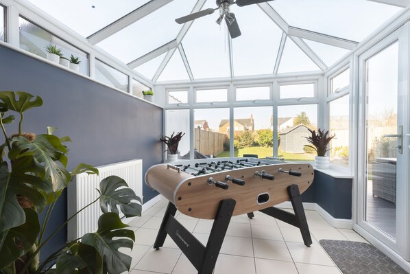 A conservatory with a foosball table, large windows, and potted plants.