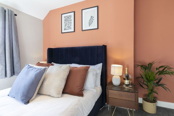 Una habitación de hotel con cama, almohadas, una mesita de noche con lámpara, una botella de champán y una planta en maceta.