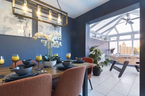 Un coin repas avec une table en bois dressée pour quatre personnes, entourée de chaises. Une grande fenêtre offre une vue sur l’extérieur, et on y trouve également un ventilateur de plafond et un luminaire fixé au mur.