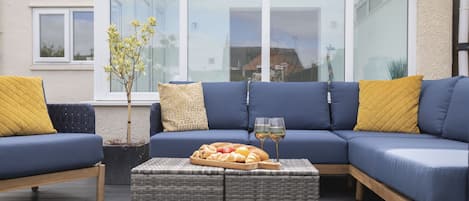 A modern outdoor seating area with a blue sofa, yellow cushions, and a wicker table with a tray of bread and drinks.