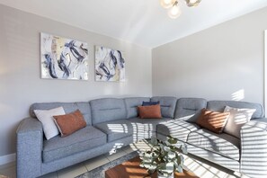 A modern living room with a grey sectional sofa, abstract wall art, and a coffee table with a plant. - 4-Bed Home with Hot Tub near Gloucester City (Gloucestershire)