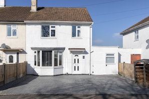 Une maison blanche à deux étages, avec un toit en tuiles, une porte d’entrée et un petit jardin devant.