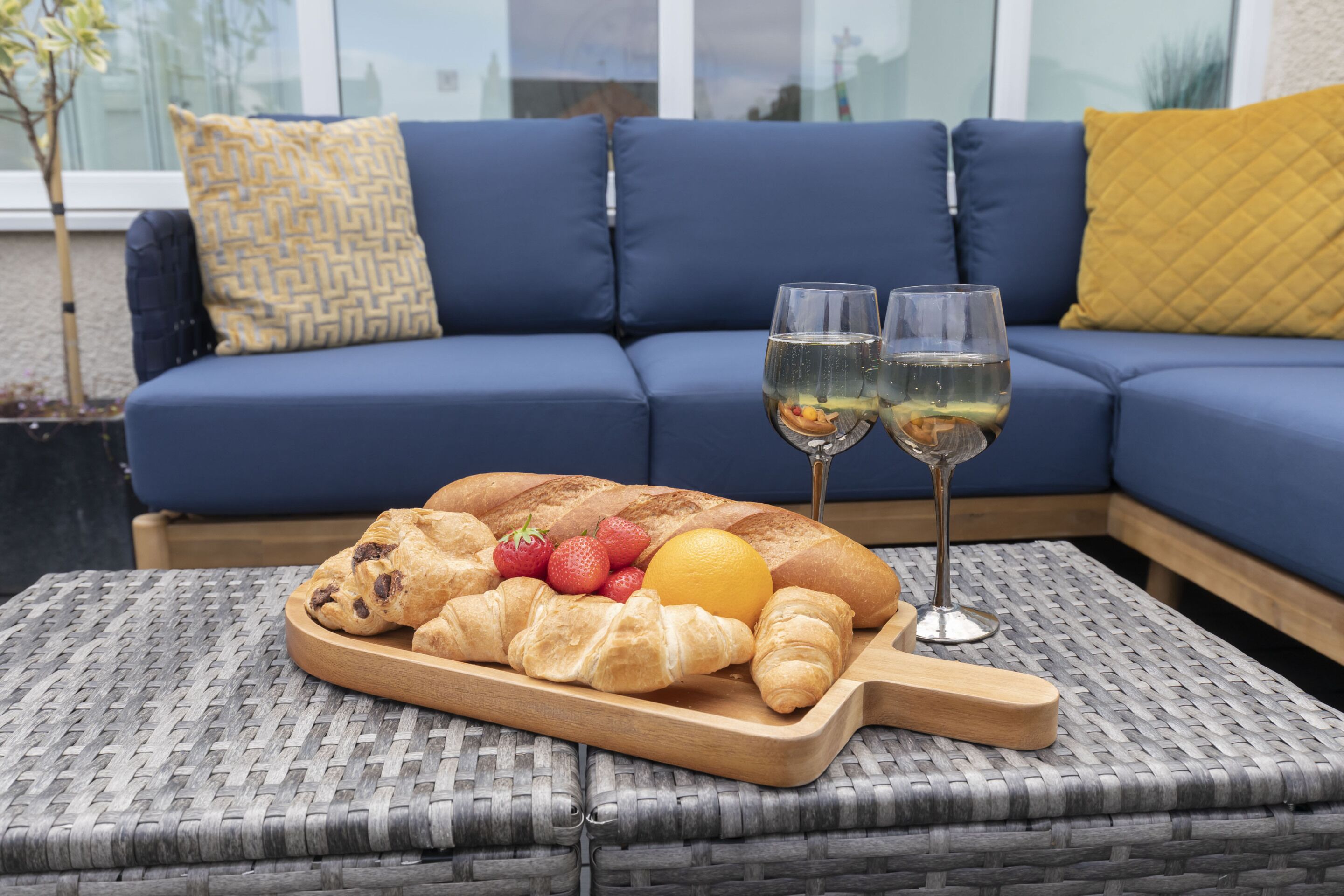 A dining setup with a wooden board of croissants, strawberries, and an orange on a woven table, with a blue sofa and cushions in the background.