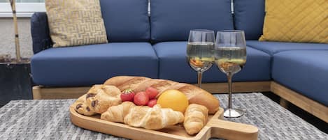 A dining setup with a wooden board of croissants, strawberries, and an orange on a woven table, with a blue sofa and cushions in the background.