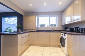 A compact kitchen with a washing machine, a sink, and a window with potted plants. - 4-Bed Home with Hot Tub near Gloucester City (Gloucestershire)
