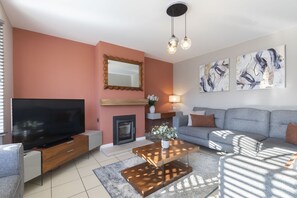 A modern living room with a flat-screen TV, a fireplace, a sofa, and abstract wall art. - 4-Bed Home with Hot Tub near Gloucester City (Gloucestershire)
