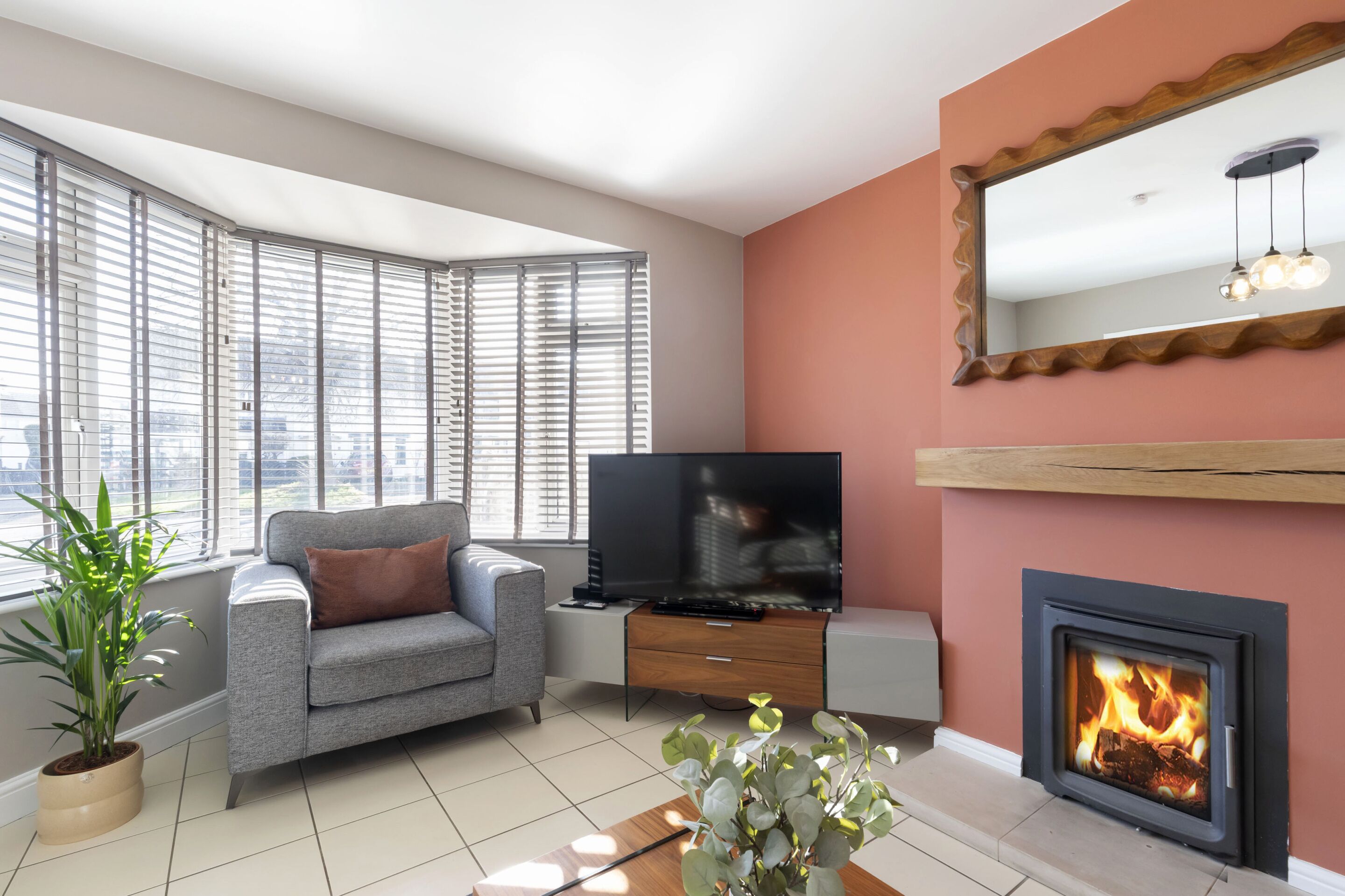 A modern living room with a grey sofa, a flat-screen TV, a fireplace, and large windows with blinds.