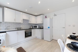 A modern kitchen with white cabinets, a microwave, a refrigerator, and a dining area with a table and chairs. - Modern 4 Bedroom 4 Bathroom Home in Cheltenham (Cheltenham)