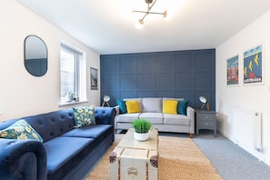 A modern living room with a blue sofa, a grey sofa, a small table with a plant, and a suitcase.