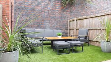 A patio with a wooden table and grey sofa, surrounded by potted plants.