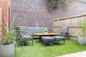 A patio with a wooden table and grey sofa, surrounded by potted plants.
