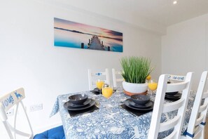 A dining area with a table set for two, a potted plant, and a sunset painting on the wall.