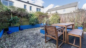 A patio with wooden furniture and a gravel ground, surrounded by a wooden fence and vegetation. - Modern & Spacious Home in Central Cheltenham (Gloucestershire)