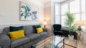 A modern living room with a grey sofa, yellow and green cushions, a glass coffee table, a large abstract painting, a green armchair, a potted plant, and a radiator. - Stunning 5 Bed Regency Home (Gloucestershire)