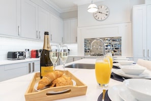 A modern kitchen with white cabinets, a wooden breakfast tray with croissants and a bottle of Valdo Prosecco, and two glasses of orange juice.