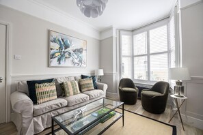 En moderne stue med et sofabord i glas, en beige sofa med mønstrede puder, en mørk lænestol og et stort abstrakt maleri på væggen.
