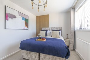 A bedroom with a large bed, a bedside table, a lamp, a window with blinds, a wall-mounted light fixture, and a piece of abstract art. - Stunning Five Bed Detached Home with Hot Tub (Gloucestershire)
