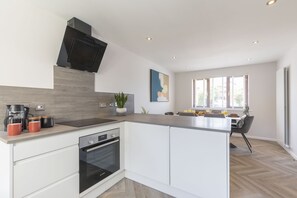 A modern kitchen with a built-in oven, a microwave, and a coffee maker. There is a dining area with a table and chairs, and a window with blinds.