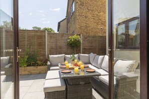 Un patio al aire libre con una mesa de comedor para cuatro personas, rodeado de muebles de mimbre y cojines.