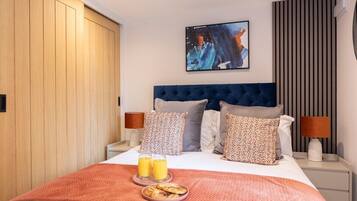 A hotel room with a bed, two bedside lamps, a painting on the wall, and a breakfast tray with two glasses of orange juice and a plate of pastries.