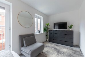 A modern bedroom with a grey sofa, a large mirror, a TV on a black dresser, and a potted plant.