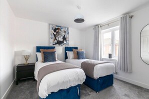 Une chambre avec deux lits, une grande fenêtre avec des rideaux, un luminaire au plafond et un tableau accroché au mur.