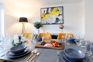 Un comedor con una mesa dispuesta para cuatro personas, con un centro de mesa que incluye pan, fruta y queso, rodeado de platos y cubiertos.