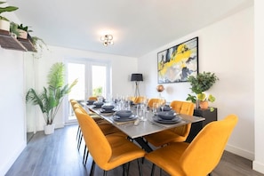 A dining area with a long table set for six, surrounded by four orange chairs. There are potted plants on the left side, a painting on the wall, and a lamp on the table.