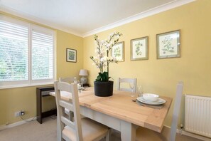 A dining area with a wooden table, white chairs, a potted plant, and framed botanical prints on the wall.