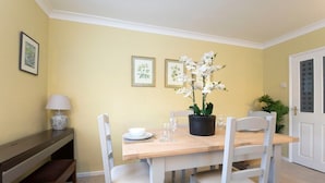 A dining area with a wooden table and white chairs, a potted orchid, and framed botanical prints on the wall.