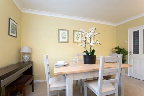 A dining area with a wooden table and white chairs, a potted orchid, and framed botanical prints on the wall.