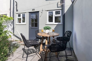 A small patio with a round wooden table and four black wicker chairs. There are two potted plants on the table, and two glasses of orange juice.