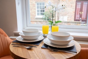 A breakfast setting with two white bowls, two plates, and two glasses of orange juice on a wooden table by a window with a view of a brick building.