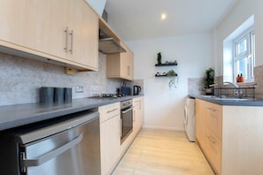 A modern kitchen with light wood cabinets, stainless steel appliances, and a built-in oven. There are shelves with plants and a window providing natural light. - Beautiful Bath Road Retreat with Log Burner (Gloucestershire)