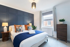 A neatly arranged bedroom with a bed, bedside lamp, dresser, and a window with curtains.