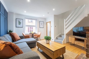 A modern living room with a sofa, coffee table, and TV stand. There is a staircase to the right and a window with blinds.