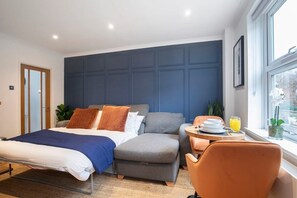 A modern hotel room with a large bed, a grey sofa, an orange armchair, and a small dining table with breakfast items.