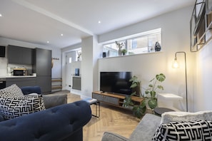 A modern living room with a large flat-screen TV, comfortable seating, a coffee table, and a dining area with a kitchen in the background.