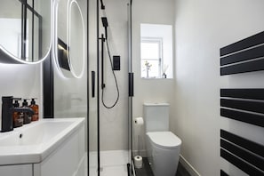 A modern bathroom with a glass-enclosed shower, a white sink with soap dispensers, a toilet, and a mirror with illuminated edges.