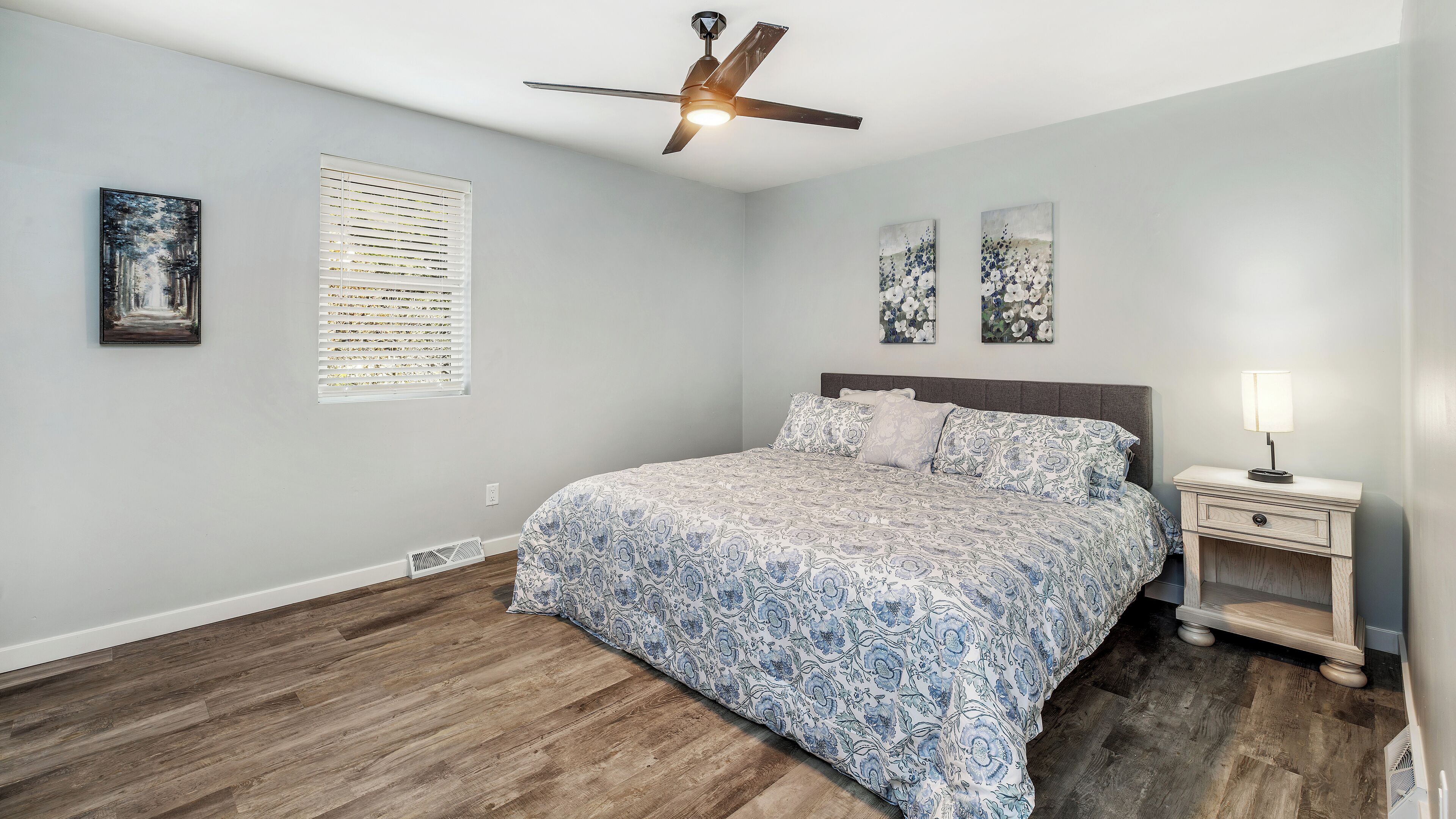 A bedroom with a bed, a nightstand, a ceiling fan, and artwork on the walls.