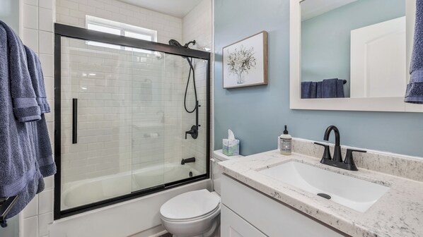 A modern bathroom with a glass-enclosed shower, a white toilet, a sink with a marble countertop, and a framed picture on the wall. - NEW! 3 bed, 1.5 Bath, Games, WIFI (Escanaba)